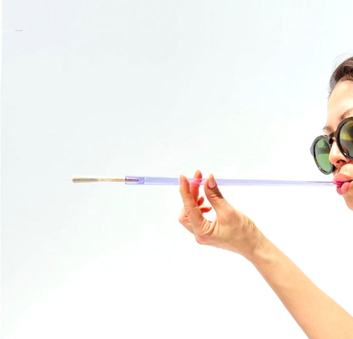 A woman holding a joint holder to her lips to demonstrate scale, it's  a 12 inch extra long Cruella joint holder, made from transparent purple glass